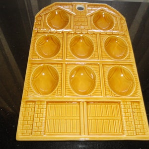 May include: A ceramic egg holder in a barn shape, featuring nine egg-shaped indentations. The holder is a warm, golden-yellow color with a textured surface, and includes a barn door design at the bottom. It has a hole at the top for hanging.