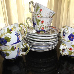 May include: A set of white ceramic teacups and saucers with floral designs. The cups and saucers have a scalloped edge and are decorated with blue, red, and green flowers. The teacups have a unique handle design.