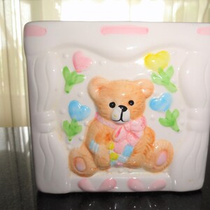 May include: A white ceramic planter with a pink trim features a brown teddy bear with a pink bow and a blue and yellow plaid blanket. The planter is decorated with pink, blue, and yellow hearts and green leaves.