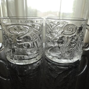 May include: Two clear glass mugs with raised images of Batman and the Joker. The mugs are empty and sitting on a black surface.