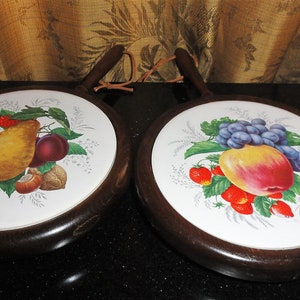 May include: Two round wooden hot pads with a white ceramic center. The ceramic centers feature painted images of fruit. One pad features a pear, plums, raspberries, and nuts. The other pad features a peach, grapes, and strawberries.