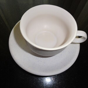May include: A light gray ceramic teacup and saucer set. The cup has a handle and sits on a matching saucer. Both pieces have a speckled pattern. The cup is empty and the set is on a dark surface.