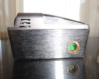 May include: A silver lighter with a brushed metal finish and a green jewel embedded in a gold ring. The lighter has a black plastic top with three air vents.