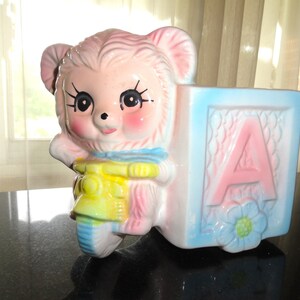 May include: A pink ceramic planter shaped like a bear holding a yellow and blue toy. The planter is attached to a blue and pink block with the letter "A" on it.