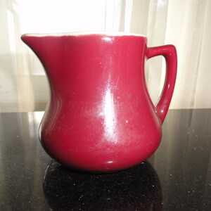 May include: A burgundy ceramic pitcher with a curved body and a handle. The pitcher has a white rim and a spout for pouring. The surface has a glossy finish. This kitchenware item is suitable for serving beverages.