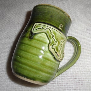 May include: Green ceramic mug with a raised outline of the state of Florida. The mug has a handle and a textured surface.