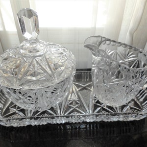 May include: A clear glass sugar bowl with a lid and a matching creamer, both with a cut glass design, are displayed on a clear glass tray.
