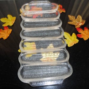 May include: A clear glass corn on the cob serving dish with six individual slots. The dish has a textured surface that resembles corn kernels.