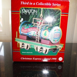 May include: A red box with a gold trim, featuring a Christmas train ornament. The ornament is a silver and red train car with a green flatbed. The train car has a green and white Christmas tree on the side and says "Happy Holidays" and "1998". The box says "Carlton Cards" and "Christmas Express - Dated 1998".