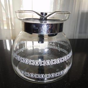 May include: A clear glass coffee carafe with a silver metal band and lid. The carafe has a rounded body and a decorative white floral pattern around the middle. The spout is designed for easy pouring.