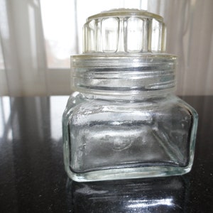 May include: A clear glass jar with a lid that has a grid pattern. The jar is square shaped and has a rounded top.
