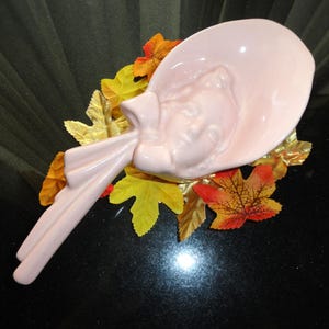 May include: A pink ceramic dish shaped like a woman's head wearing a hat. The dish is surrounded by fall leaves in shades of gold, orange, and red.