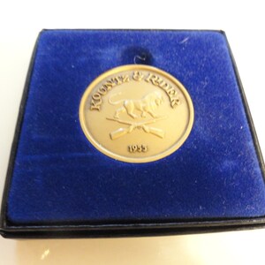 May include: A gold-colored coin with a lion and crossed rifles design. The coin is in a blue velvet box with a black border. The text "KOONTZ & RIDER" and "1935" are inscribed on the coin.