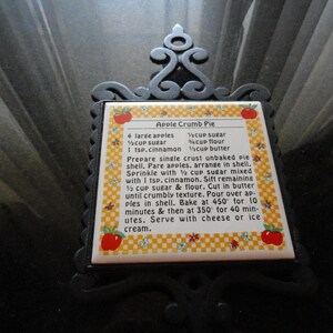May include: A white ceramic tile with a yellow and white checkered background. The tile has a red apple graphic and a recipe for Apple Crumb Pie. The recipe includes ingredients like apples, sugar, flour, and cinnamon. The tile is framed with a black metal frame.