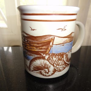 May include: A ceramic mug with a coastal design. The mug features a brown boat, seashells, and seagulls against a white background. Brown bands accent the top. The mug has a white handle.