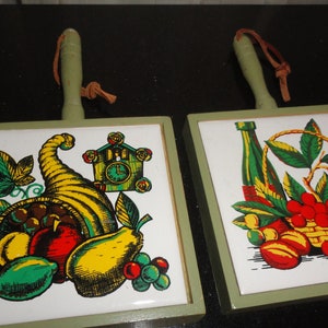 May include: Two vintage green framed ceramic trivets with wooden handles. One trivet features a cornucopia with fruit and a cuckoo clock. The other trivet features a bottle of wine, grapes, and a basket of cherries.