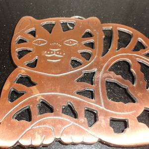 May include: A copper-colored cat-shaped ornament with cut-out details. The cat's face is visible, with eyes, nose, and whiskers. The ornament has a small loop at the top for hanging. The cat is lying down.