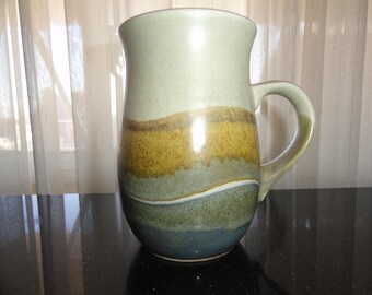 May include: A ceramic mug with a light brown, green, and blue glaze. The mug has a rounded body and a handle.