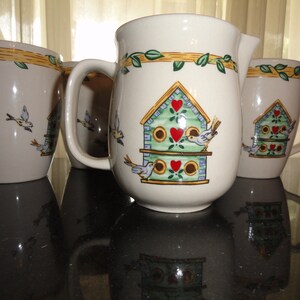May include: Set of three ceramic mugs and a pitcher with a birdhouse design. The mugs and pitcher are white with green, yellow, and brown accents. The birdhouse features a green roof, red hearts, and brown accents. The birds are white with black eyes and yellow beaks.