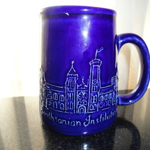 May include: A blue ceramic mug with a white embossed image of the Smithsonian Institution building. The mug has a handle and the text "Smithsonian Institution" is visible on the bottom.
