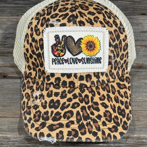 May include: A baseball cap with a leopard print design and a mesh back. The front of the cap has a patch with a peace sign, a heart, a sunflower, and the words "PEACE LOVE SUNSHINE". The cap has a distressed appearance.