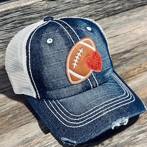 Puede incluir: Una gorra de camionero de mezclilla azul con una malla blanca en la parte posterior. La gorra tiene un aspecto desgastado y presenta un balón de fútbol de cuero marrón con un corazón rojo de brillantina.