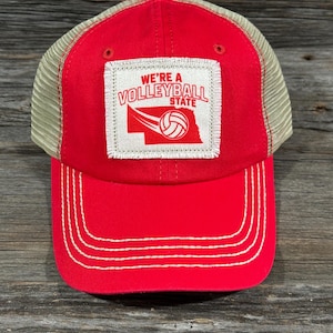 May include: Red and beige mesh-back baseball cap with a curved peak. The front displays a white patch with the text "WE'RE A VOLLEYBALL STATE" and a volleyball graphic. The cap has white stitching and a red button on top.