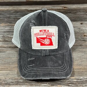 Puede incluir: Gorra de camionero negra desgastada con parte trasera de malla blanca. El parche frontal muestra el texto "WE'RE A VOLLEYBALL STATE" en rojo, junto con un gráfico de voleibol. La gorra tiene una visera curva y costuras visibles.