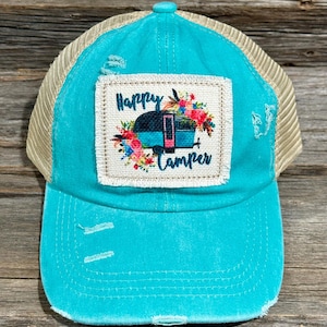 May include: A turquoise baseball cap with a beige mesh back. The front features a patch with the words "Happy Camper" above a camper illustration surrounded by flowers. The cap has a distressed look with frayed edges.