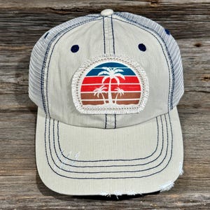 Op de afbeelding: Beige en witte trucker cap met een versleten klep. Het heeft een patch met een palmboomontwerp in rood, blauw en bruin. De cap heeft een mesh rug en marineblauwe stiksels.