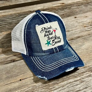 Op de afbeelding: Een blauwe denim trucker-pet met een wit mesh-achterpaneel. De pet heeft een distressed look en heeft een patch met de tekst "Drink in my hand toes in the sand" en een rode cocktailglas-afbeelding.