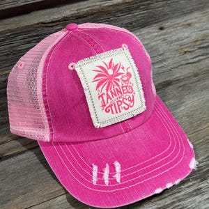 May include: A distressed pink trucker hat with a mesh back and a curved brim. The hat features a square patch with a palm tree graphic and the words "Tanned and Tipsy". The hat has a faded, worn look.