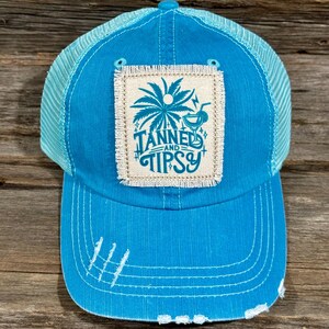 May include: A turquoise trucker hat with a mesh back and a curved brim. The front features a square patch with a palm tree and the words "Tanned and Tipsy". The hat has distressed details and white stitching.