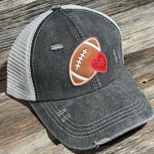 Puede incluir: Una gorra de camionero negra y gris desgastada con una pelota de fútbol marrón y blanca y un corazón rojo en la parte delantera.