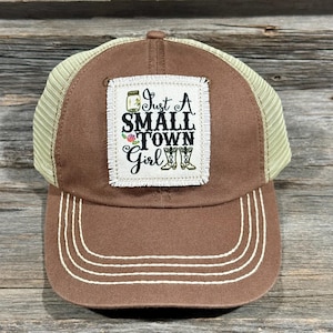 May include: Brown and tan trucker hat with a patch that says "Just A Small Town Girl" with a pair of boots and flowers.