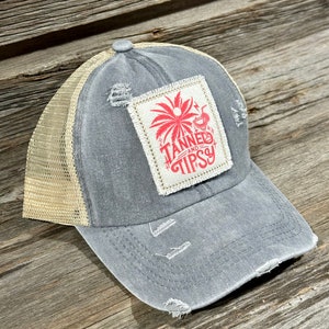 May include: A grey and beige distressed trucker hat with a patch that says "Tanned and Tipsy" with a palm tree and a cocktail glass graphic.