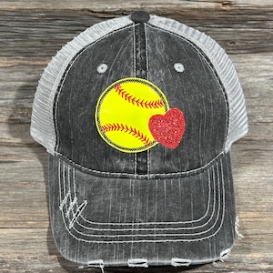 Peut inclure: Casquette de baseball noire délavée avec un dos en filet blanc. L'avant présente un motif de softball jaune avec des coutures rouges et un cœur rouge à paillettes. La casquette a une visière incurvée et un bouton sur le dessus.