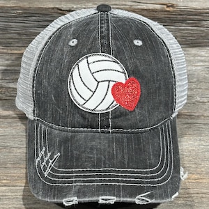 Peut inclure: Casquette de camionneur noire et grise vieillie avec un ballon de volley-ball blanc et un cœur rouge. Le chapeau a un dos en filet et une visière incurvée avec des coutures blanches. Le ballon de volley-ball a des coutures noires.