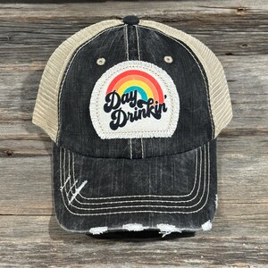 May include: Black trucker hat with a distressed finish and a beige mesh back. The front features a patch with a rainbow and the words "Day Drinkin'" in a playful font. The hat has a curved brim and white stitching.