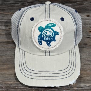 Pode incluir: Boné de beisebol creme com parte traseira em malha e aba desgastada. O boné apresenta um patch circular com um desenho de tartaruga marinha azul e as palavras "Beach Vibes". O boné tem costuras e detalhes em azul marinho.