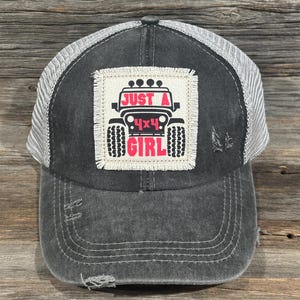 Puede incluir: Gorra de camionero negra desgastada con la parte trasera de malla blanca. La parte delantera presenta un parche cuadrado con un gráfico de Jeep y el texto "JUST A 4x4 GIRL" en rosa y negro.