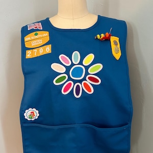 May include: A blue Girl Scout vest with a flower patch, a Girl Scouts USA patch, a Central Indiana patch, and a number 2796 patch. The vest also has a yellow and red pin and a yellow patch with a gold star.