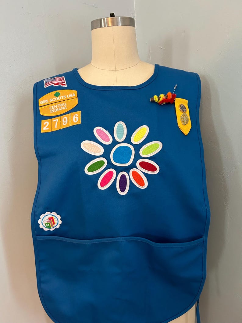 May include: A blue Girl Scout vest with a flower patch, a Girl Scouts USA patch, a Central Indiana patch, and a number 2796 patch. The vest also has a yellow and red pin and a yellow patch with a gold star.