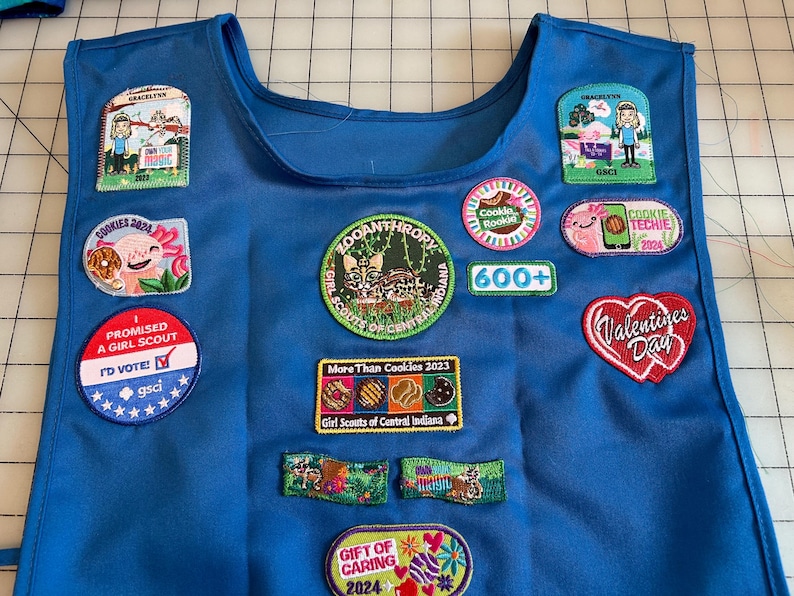 May include: A blue Girl Scout vest with various patches, including "I Promised a Girl Scout", "Cookies 2024", "Zooanthropy", "More Than Cookies 2023", "Gift of Caring 2024", "Valentines Day", "Cookie Rookie", "Cookie Techie 2024", and "600+".