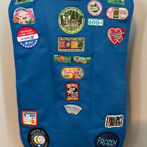 May include: A blue Girl Scout vest with various patches, including "Zooanthropy", "More Than Cookies 2023", "Gift of Caring", "Animal Shelter", "Camp Ada", "Eclipse Day 2024", "First Meeting", "2024 Camp Ada Rocks", "Frozen", and "Halloween Party".