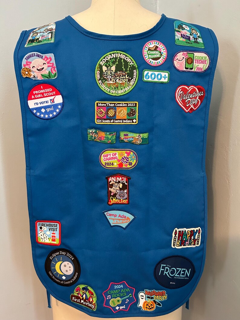 May include: A blue Girl Scout vest with various patches, including "Zooanthropy", "More Than Cookies 2023", "Gift of Caring", "Animal Shelter", "Camp Ada", "Eclipse Day 2024", "First Meeting", "2024 Camp Ada Rocks", "Frozen", and "Halloween Party".