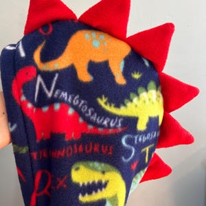 May include: A blue fleece hooded sweatshirt with a dinosaur print and red spikes on the hood. The dinosaurs are different colors and have names like Stegosaurus and Triceratops.
