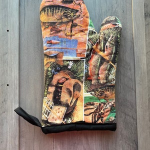 May include: A colorful oven mitt with a fishing theme. The mitt features images of fish, fishing lures, and other fishing-related items. The mitt is black with a red trim.