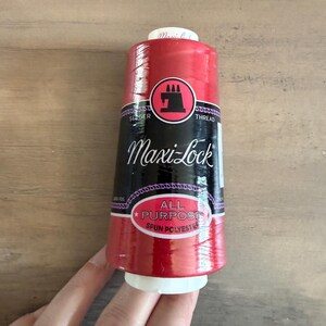 May include: A red Maxi-Lock all-purpose spun polyester thread spool. The spool has a black label with white text and a red and black circular logo. The spool is held by a hand.