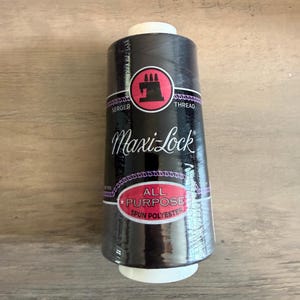 May include: A black spool of Maxi-Lock serger thread. The label features a pink circle with a sewing machine graphic and the words "SERGER THREAD". The bottom label reads "ALL PURPOSE SPUN POLYESTER".